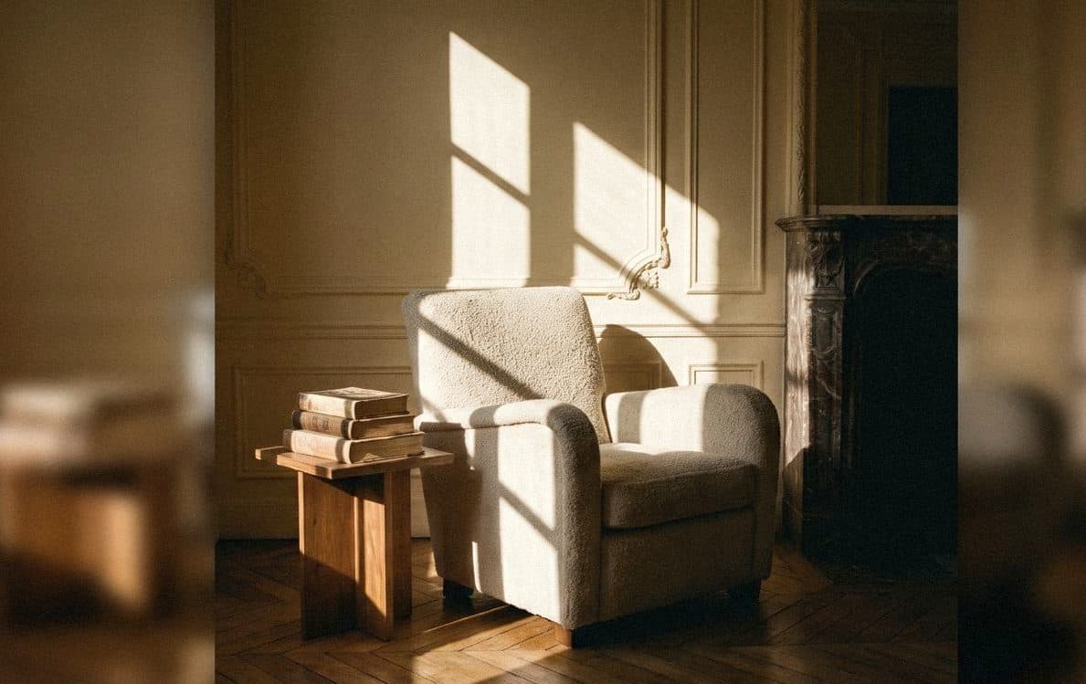 Cream bouclé armchair against a panelled cream wall in morning sun, with a wooden side table of books and a marble fireplace