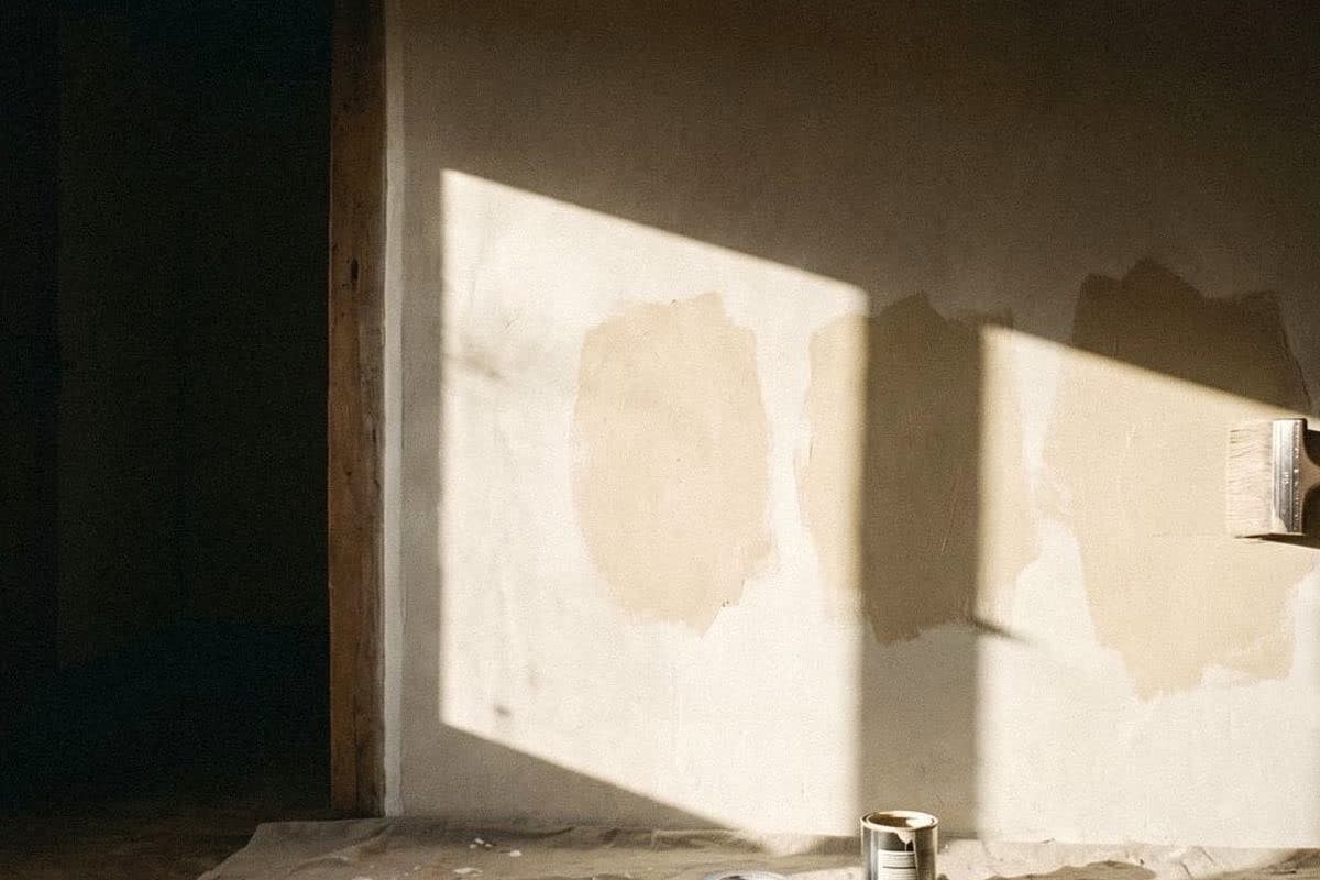 Beige test patches painted on a sunlit wall, with a paintbrush and an open tin of paint