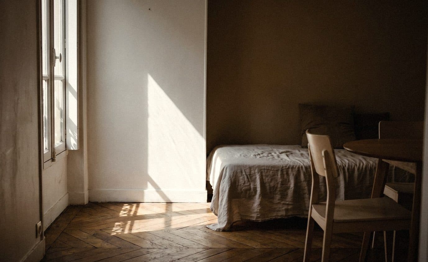 Small sunlit room with white and warm taupe walls, a linen-covered daybed and a pale wooden table and chairs