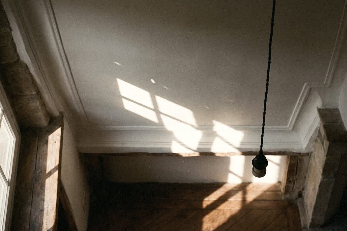 Sunlight raking across a matt white ceiling with plaster cornice, a bare black pendant cord hanging down