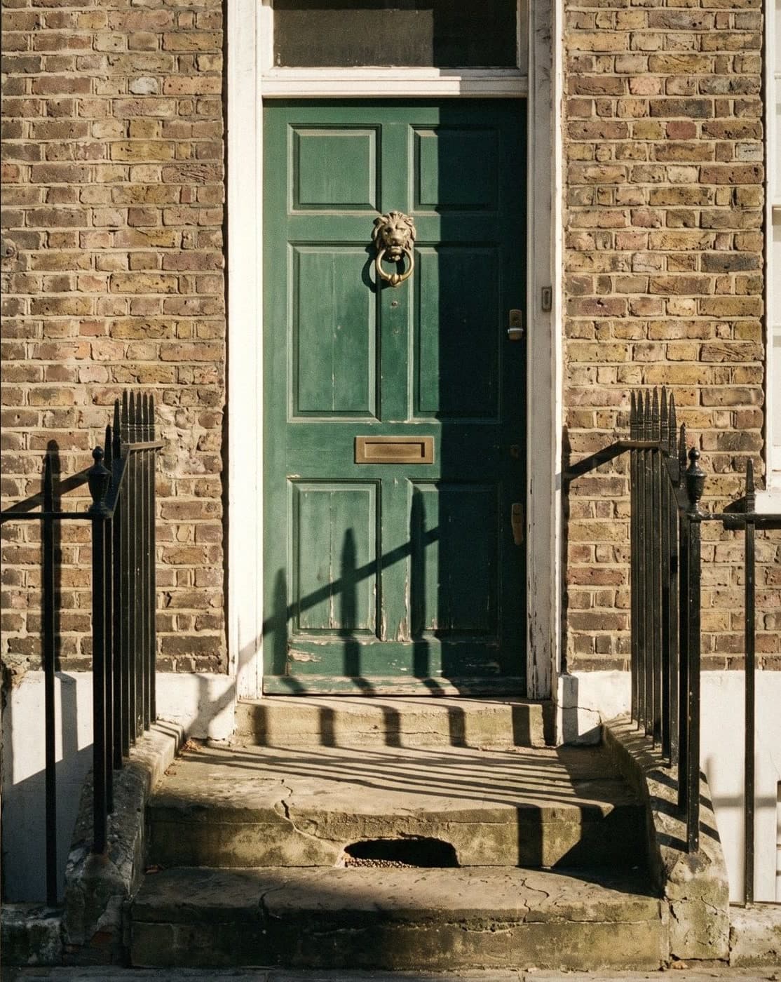 A dark green panelled front door with a brass lion knocker, set in London brick between black iron railings