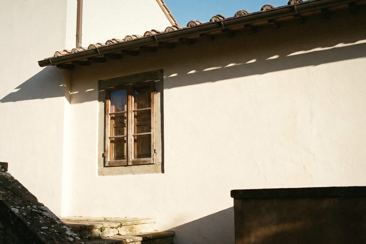 An off-white exterior wall under a terracotta-tiled eave, with a wooden window and hard sun shadows