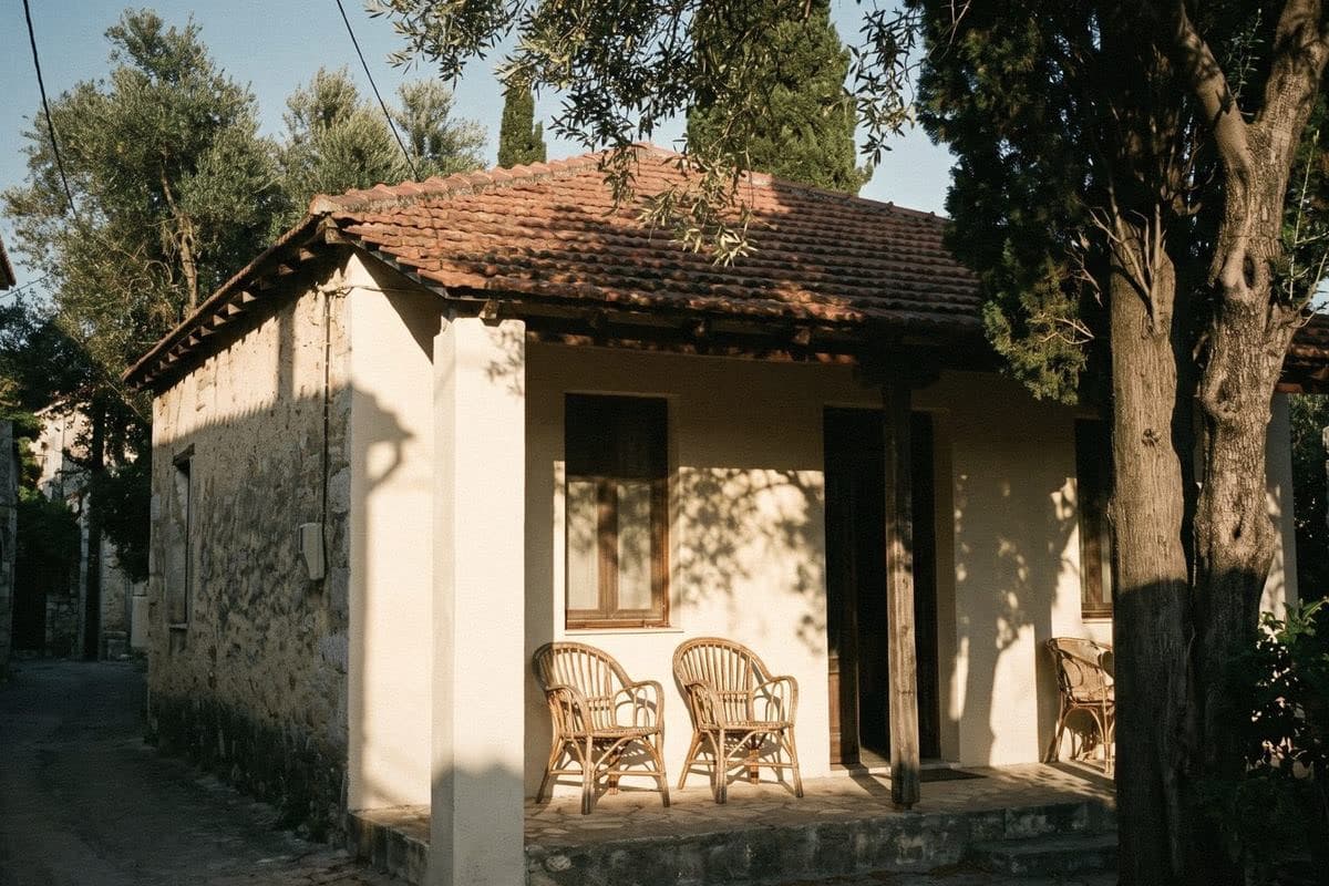 A small cream cottage with a terracotta roof, wooden windows and wicker chairs on the porch, under tall trees