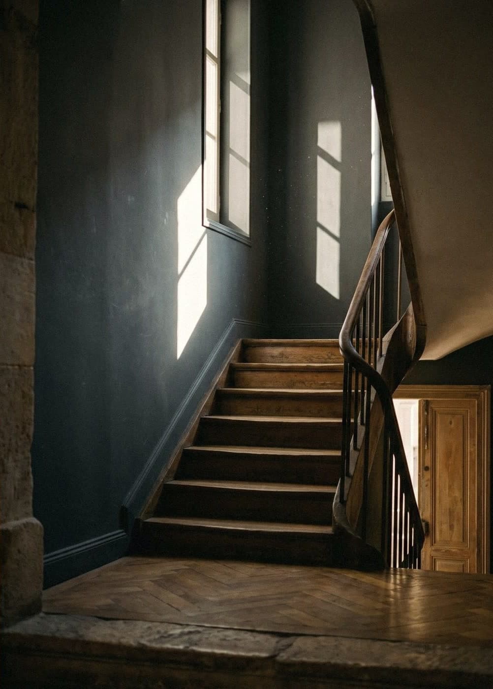 An old wooden staircase in a hall with dark blue-grey walls, sunlight from tall windows falling across them