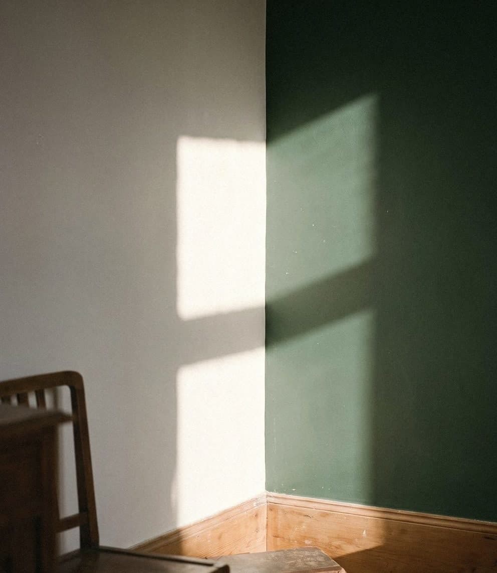 A corner where a white wall meets a deep green wall, with sun falling across both and a wooden chair