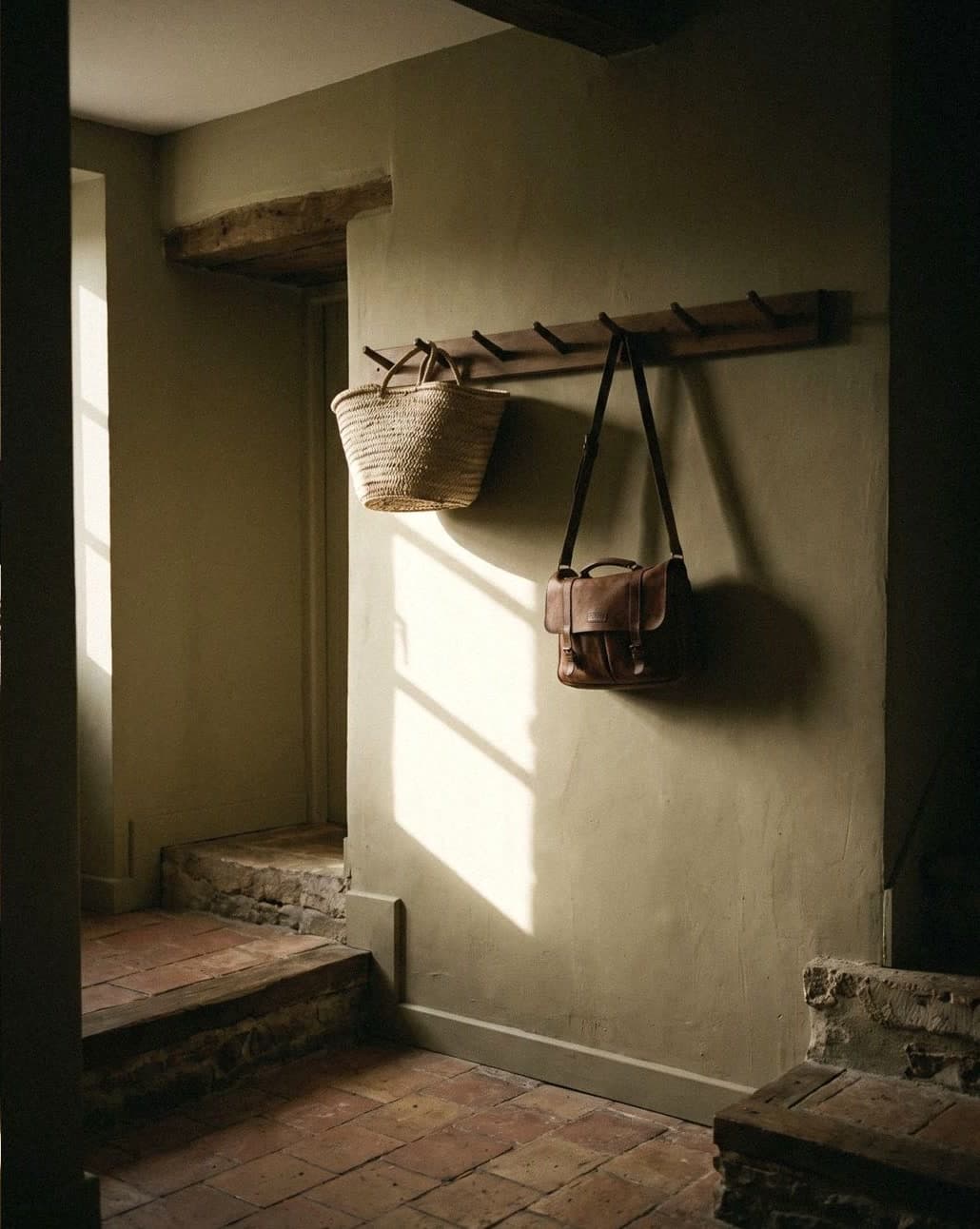 An entrance with a greenish khaki wall, a peg rail with a woven basket and leather bag, and terracotta tiles