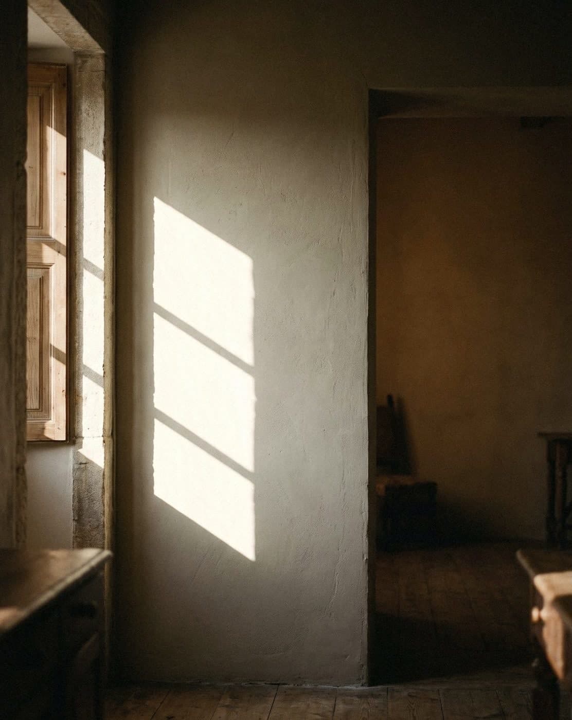 Off-white rough lime-like wall with a slanting window patch of light, beside wooden shutters and an open doorway
