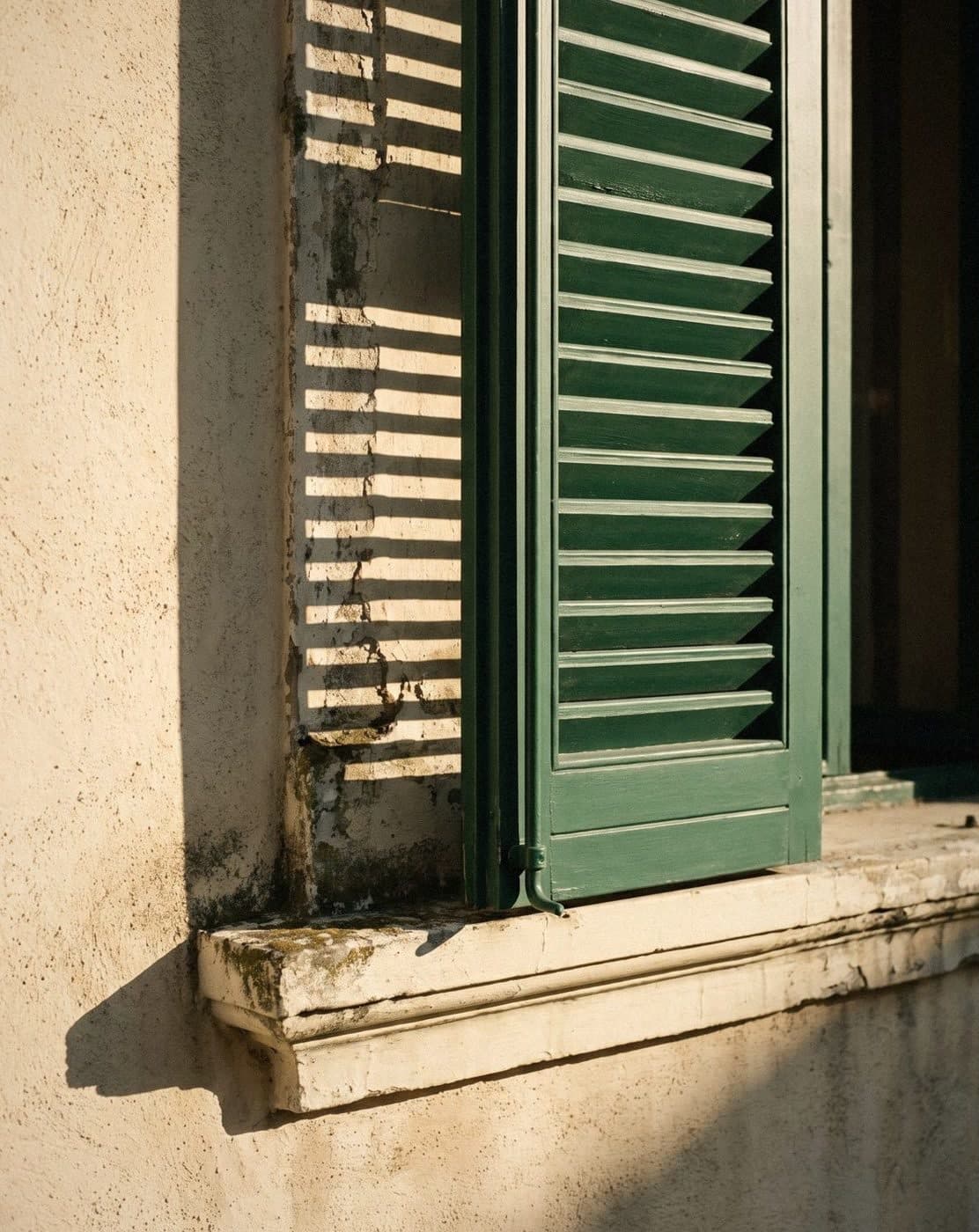 A green louvred shutter casting striped shadows across a sunlit cream rendered wall and stone sill