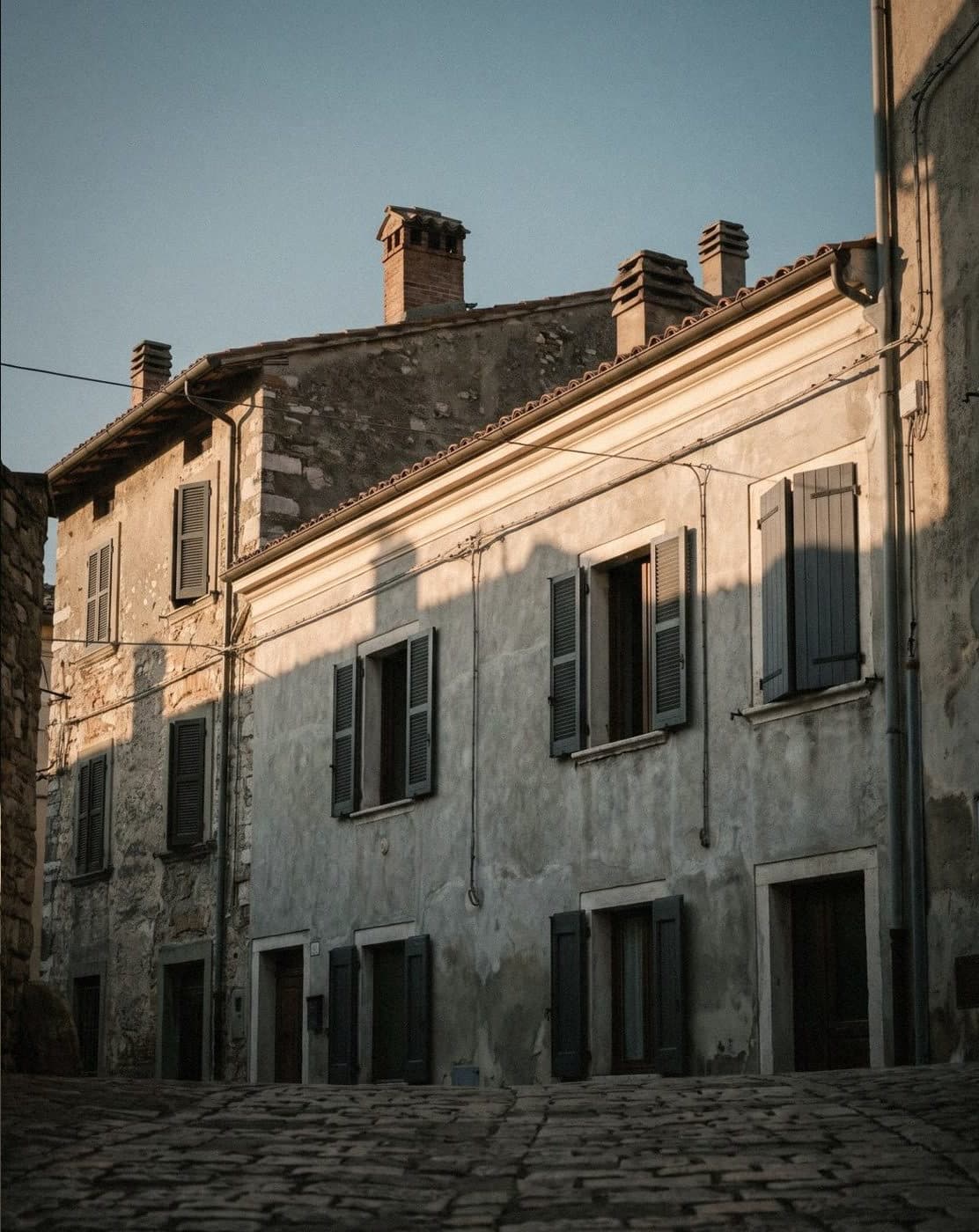 Pale grey rendered and old stone houses on a cobbled street, with dark grey shutters in late sun