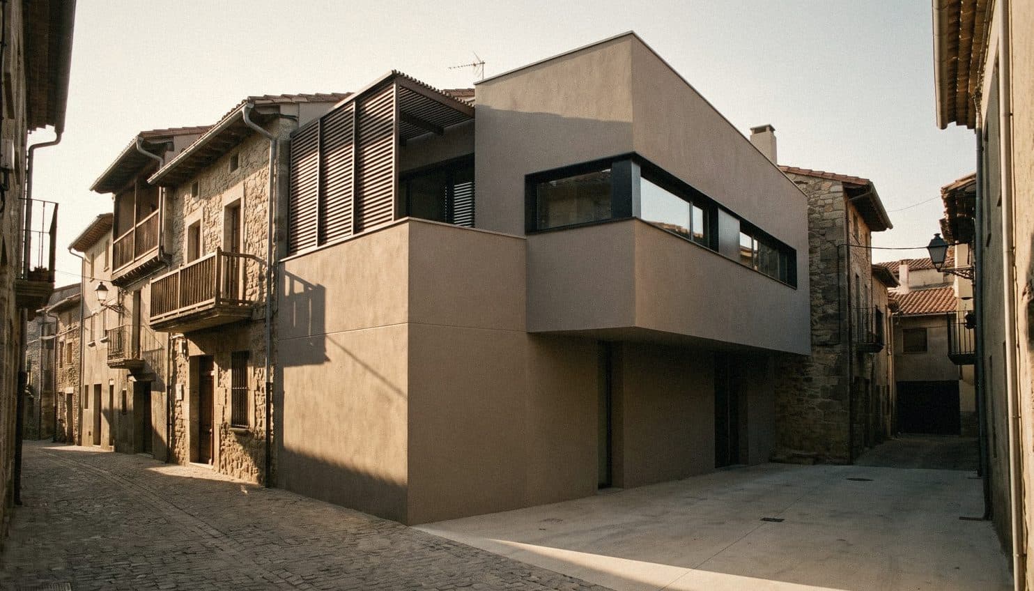 A modern taupe rendered house of stacked boxes with a long ribbon window and timber louvres among old stone houses