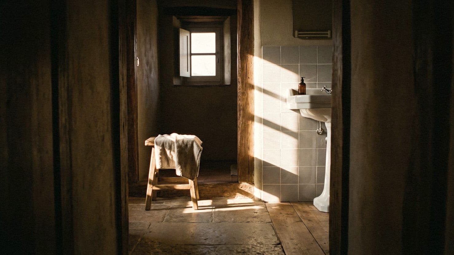 View through a doorway into a bathroom with white wall tiles in slanting sun, a pedestal basin and towel-draped stool