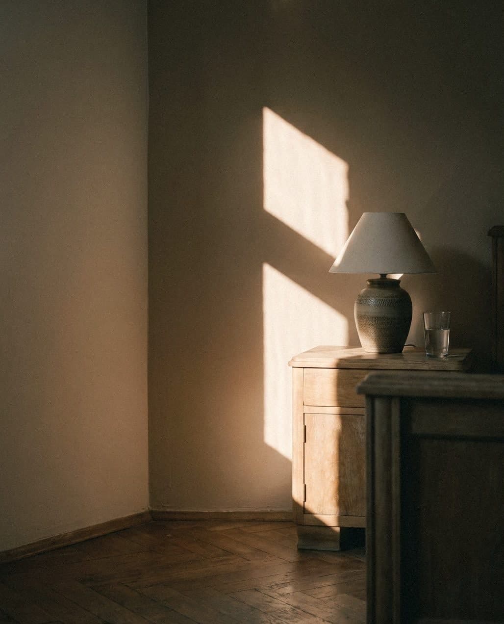 A warm beige bedroom corner with a pale wood bedside cabinet, ceramic lamp and glass of water in window-shaped sunlight