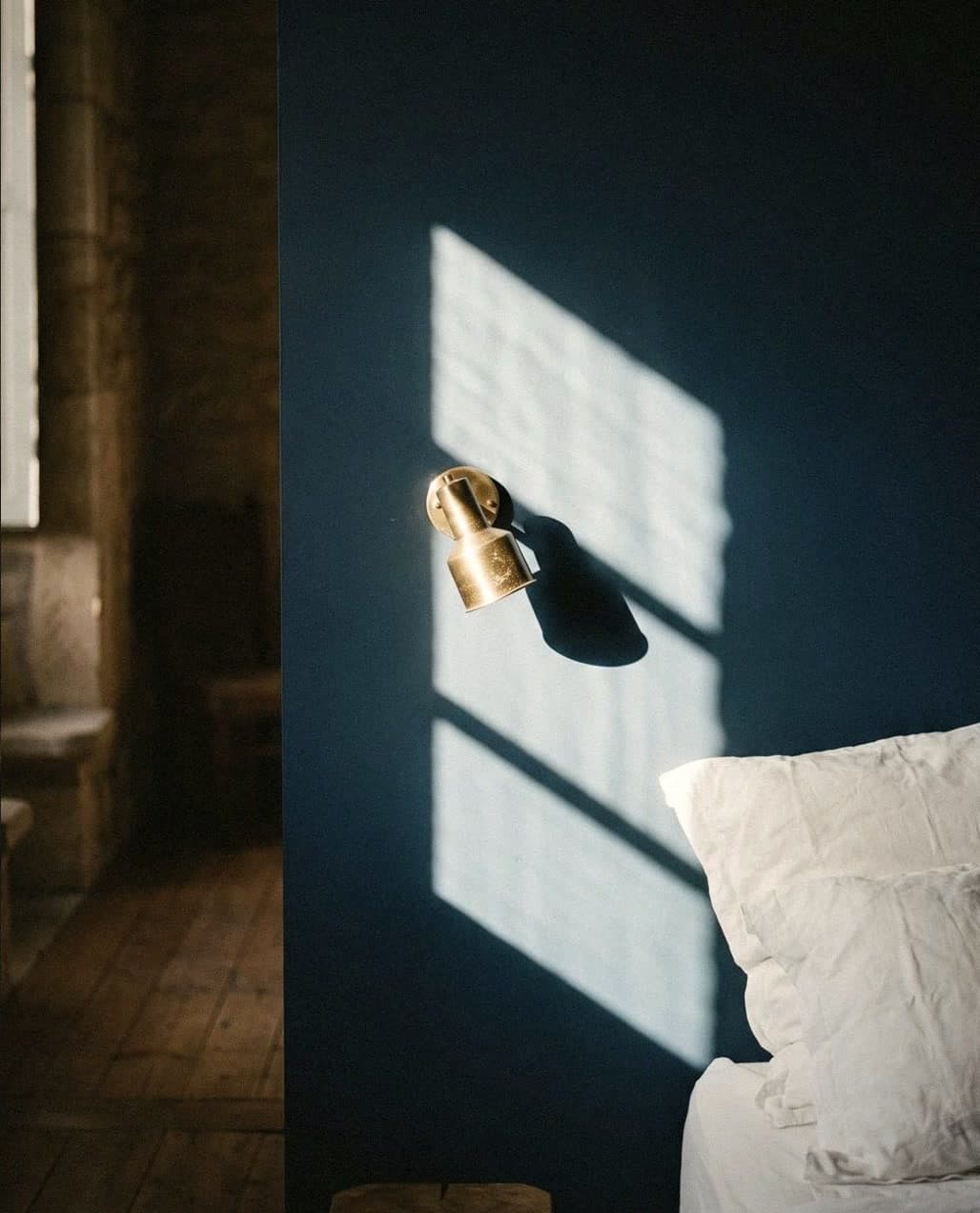 A deep navy headboard wall with a brass wall light and white linen pillows, window light falling across the paint