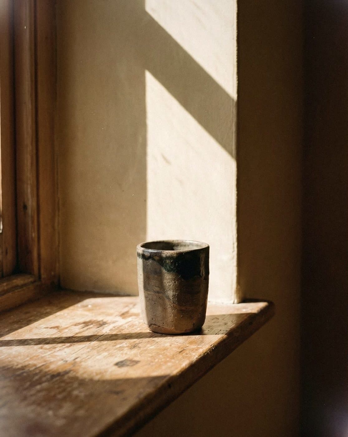 Handmade glazed stoneware cup on a worn wooden window ledge against a cream wall cut by diagonal sunlight