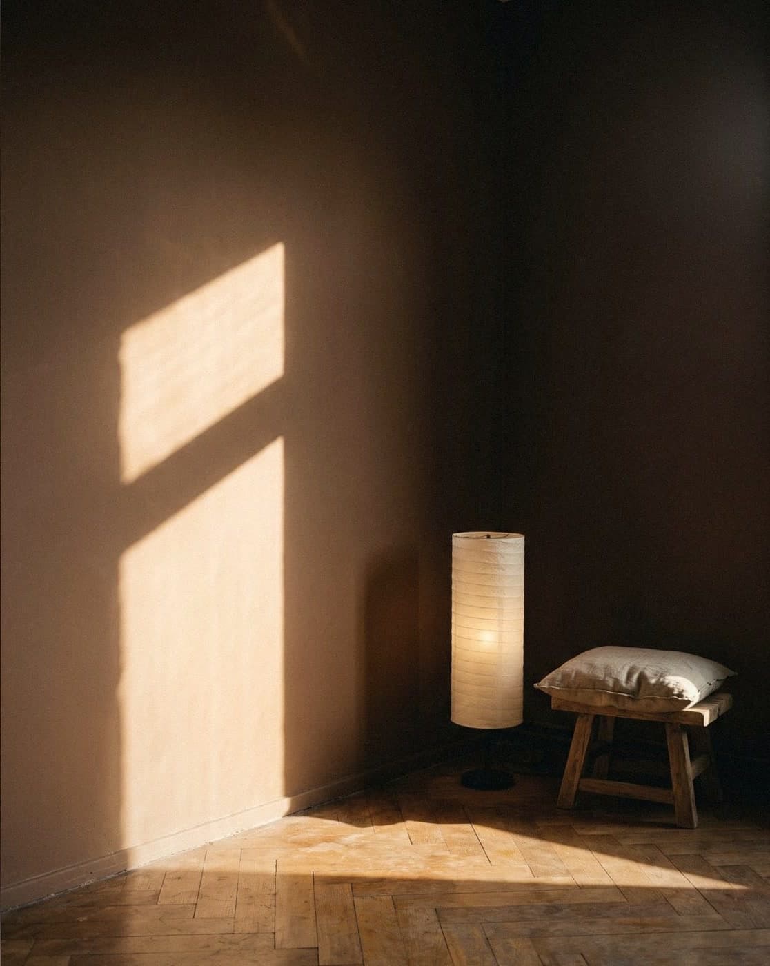 Corner with a soft pinkish-beige matt wall in window light, a tall paper lantern and a wooden stool with a linen cushion