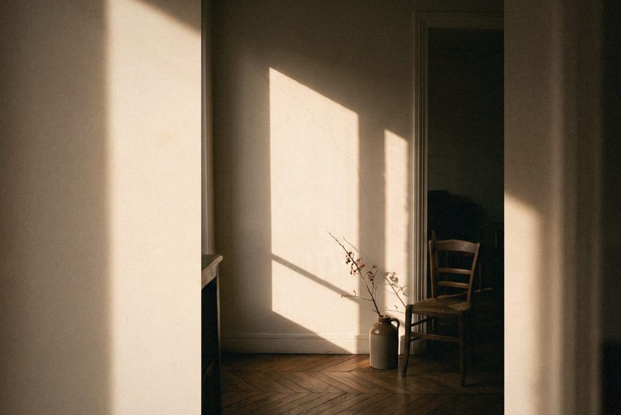 A sunlit cream wall seen through a doorway, a wooden chair and a jug of dried stems on a herringbone floor