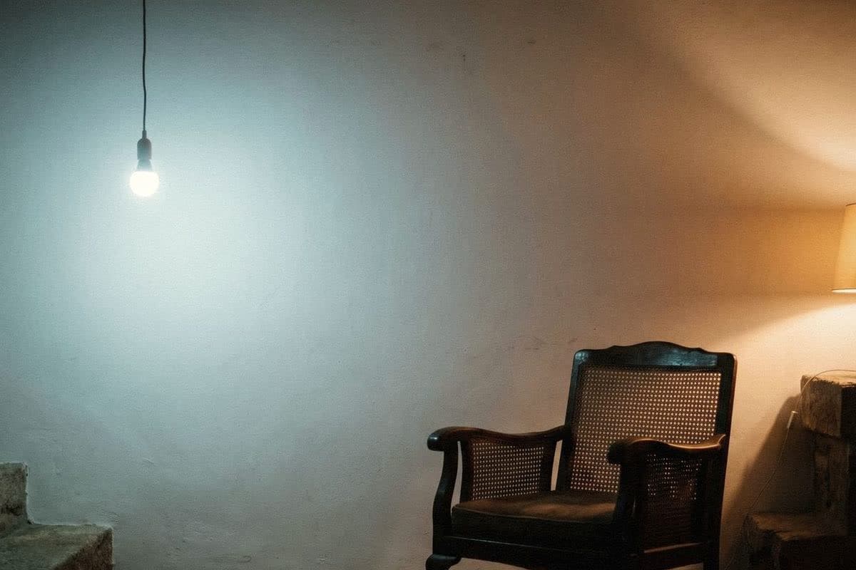 One white wall lit cool blue by a bare bulb and warm amber by a lamp, beside a cane armchair